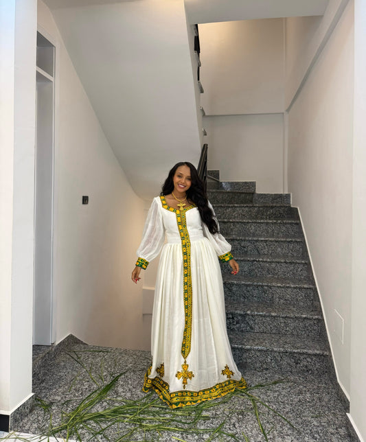 A woman wearing a long white  Ethiopia store  with yellow and green embroidered details stands on a staircase landing in a modern interior.