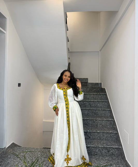 A woman wearing a long white traditional Ethiopia store with green and gold embroidery stands on a gray carpeted staircase, waving to the camera.