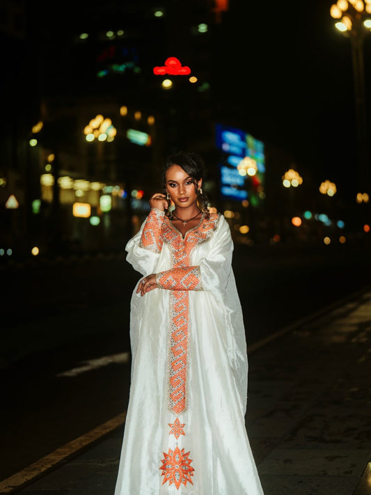 A woman wearing a white traditional long Ethiopian Clothing with orange embroidery stands on a city street at night with blurred lights in the background.