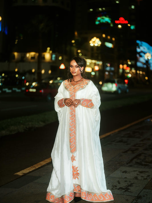 A woman wearing a white traditional long Ethiopian Clothing with orange embroidery stands on a city street at night with blurred lights in the background.