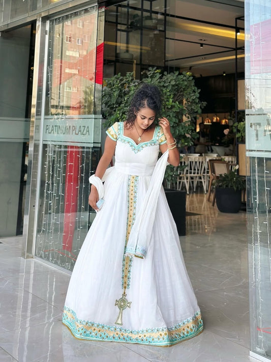 Woman in a white and green traditional Ethiopian Kemis
 standing in front of a glass door.
