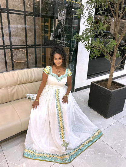Woman in a white Ethiopian kemis with green and yellow details sitting on a couch in a modern interior setting.