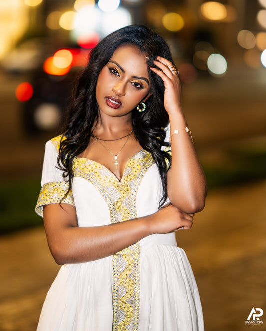 A woman posing outdoors at night wearing a white Ethiopian national dresses with yellow and silver patterned trim. The background features blurred city lights and bokeh.