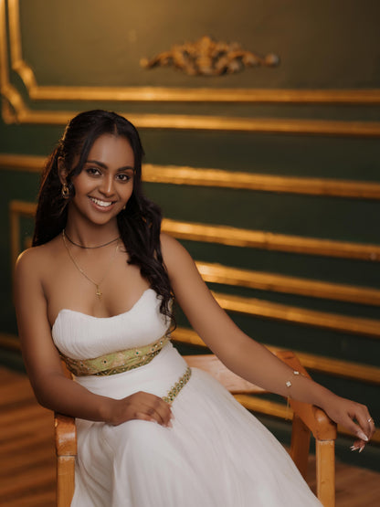 Woman in a white Habesha Kemis  with a gold belt sitting on a wooden chair against a decorative wall.