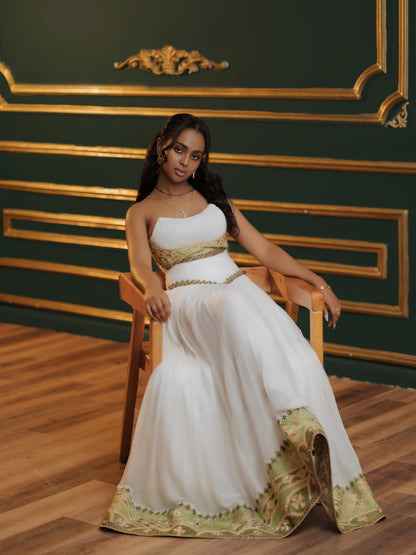 Woman in a white and gold Habesha Kemis  sitting on a wooden chair against a green wall with gold accents.