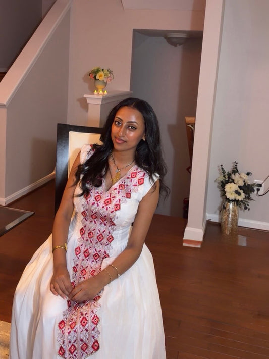 A woman wearing a sleeveless white Ethiopian Traditional Clothing with red and black geometric embroidery, sitting in an indoor home setting with wooden floors and staircase.