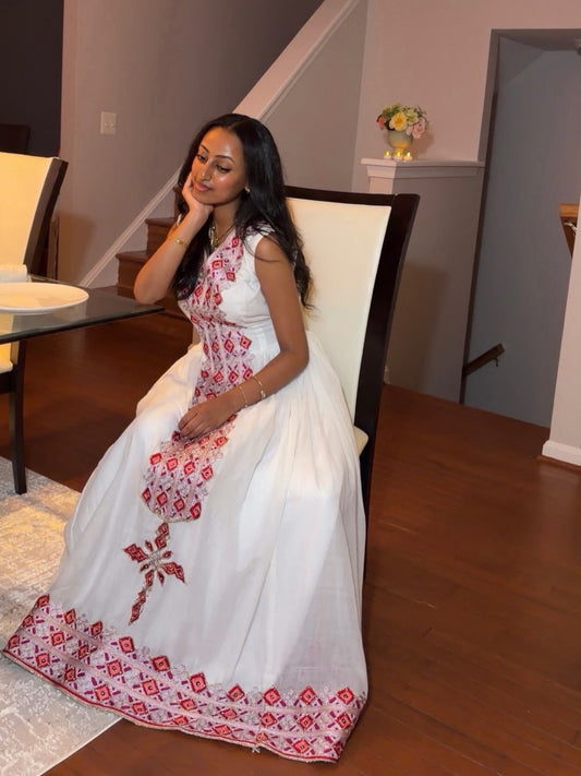 A woman wearing a white sleeveless maxi Ethiopian Traditional Clothing with red geometric embroidery, sitting on a high-backed chair in a home interior with wooden floors.