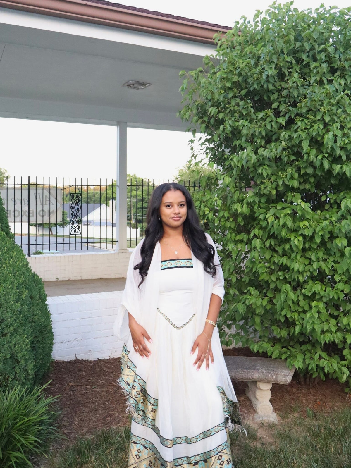 A woman wearing a white traditional-style Habesha dress  with green and gold patterned borders, standing outdoors in front of a house porch with green bushes.