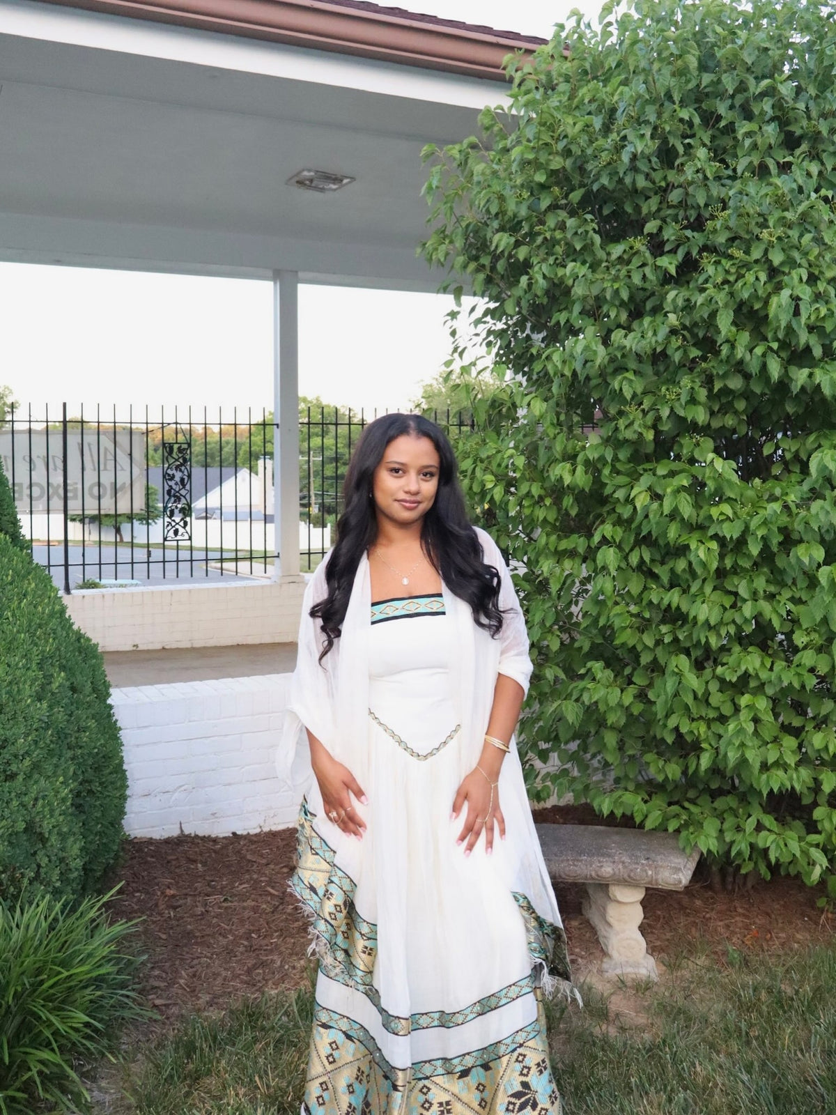 A woman wearing a white traditional Habesha dress  with green and gold border details and a matching white shawl stands outdoors in front of a house porch with green bushes.