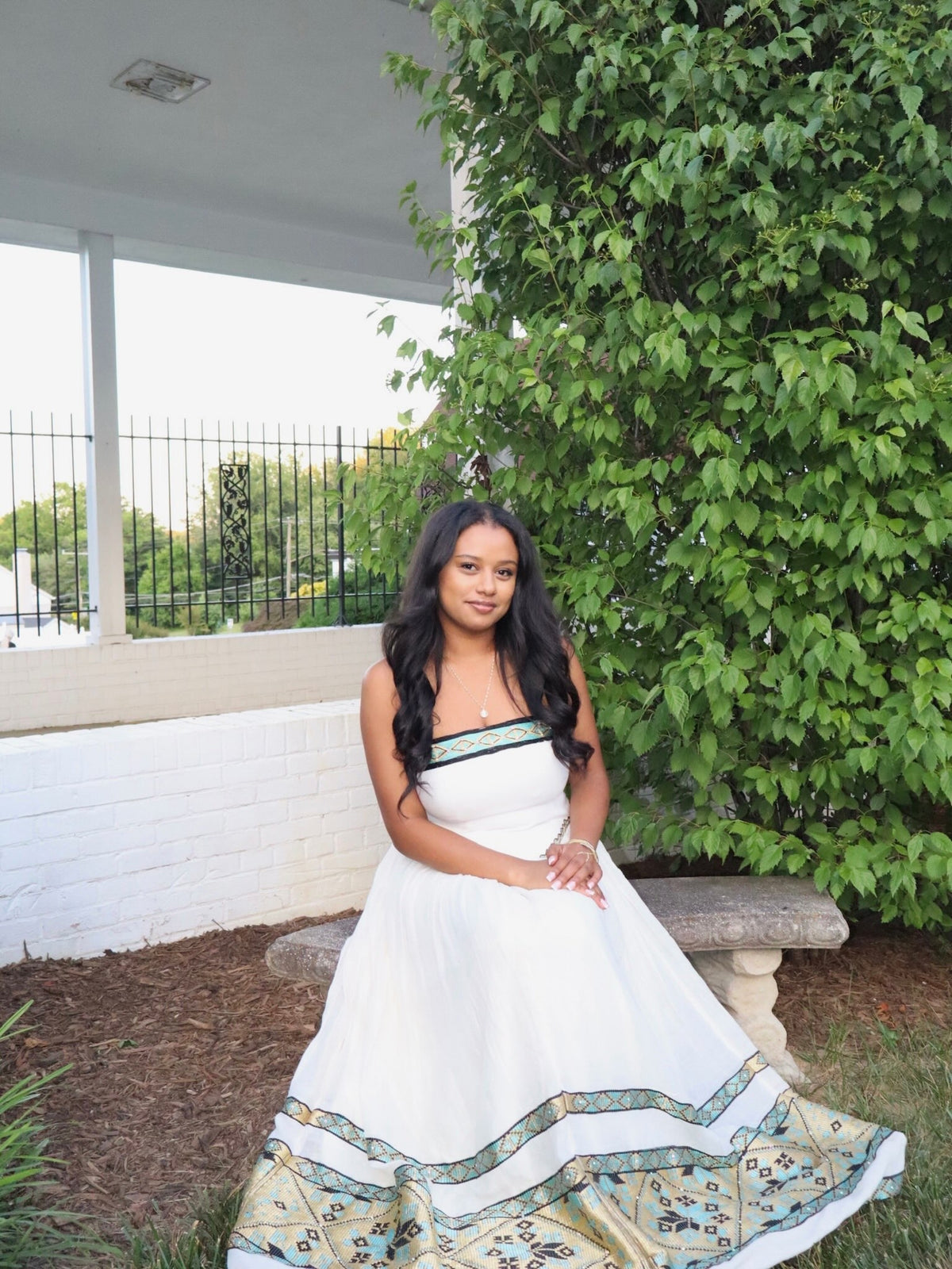 A woman wearing a white strapless Habesha dress  with green and gold patterned trim sits on a stone bench outdoors next to a leafy green tree.