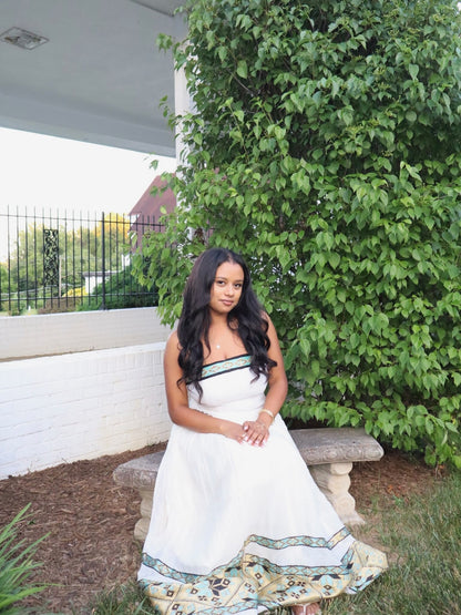 A woman with long black wavy hair sits on a stone bench outdoors, wearing a white strapless maxi Habesha dress  with green trim and a patterned hem. She is posed in front of a large green leafy bush next to a white brick wall.