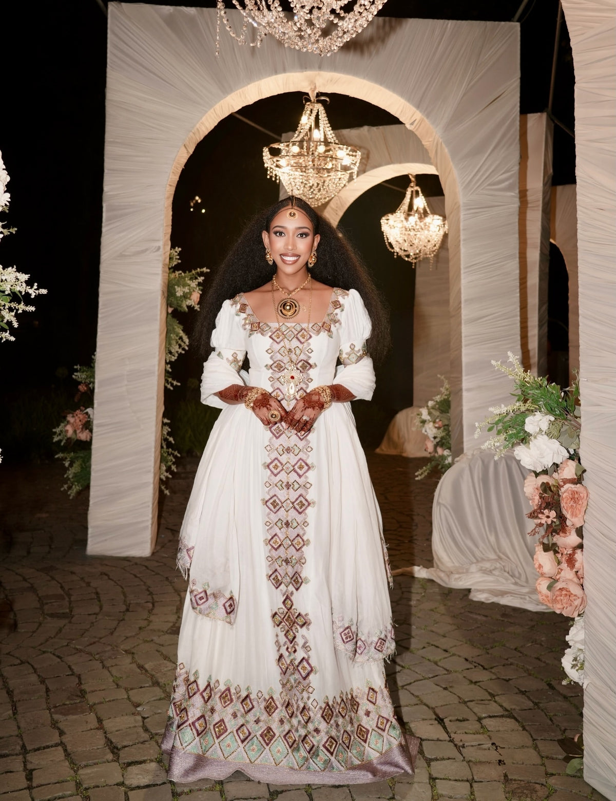 A woman stands in a decorated event space wearing a long white traditional Habesha Kemis with ornate brown and green embroidered geometric patterns. The background features draped white fabric arches, hanging chandeliers, and floral arrangements.