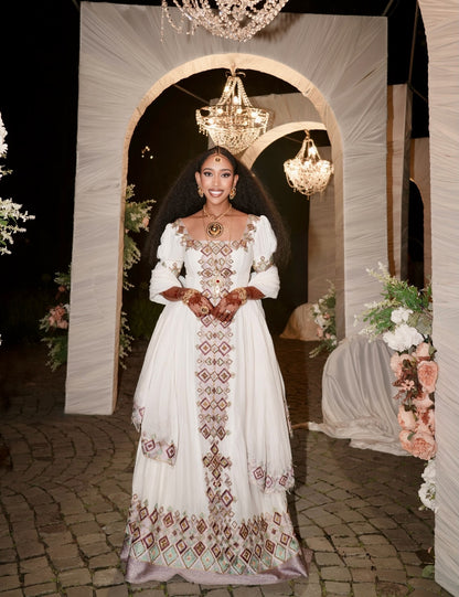 A woman stands in a decorated event space wearing a long white traditional Habesha Kemis with ornate brown and green embroidered geometric patterns. The background features draped white fabric arches, hanging chandeliers, and floral arrangements.