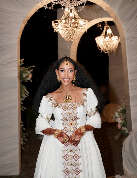 A woman stands in a decorated event space wearing a long white traditional Habesha Kemis with ornate brown and green embroidered geometric patterns. The background features draped white fabric arches, hanging chandeliers, and floral arrangements.