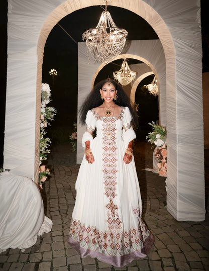 A woman stands in a decorated nighttime event setting framed by draped white arches and hanging chandeliers, wearing a long white embroidered Habesha Kemis with ornate geometric detailing and a lavender-purple border.