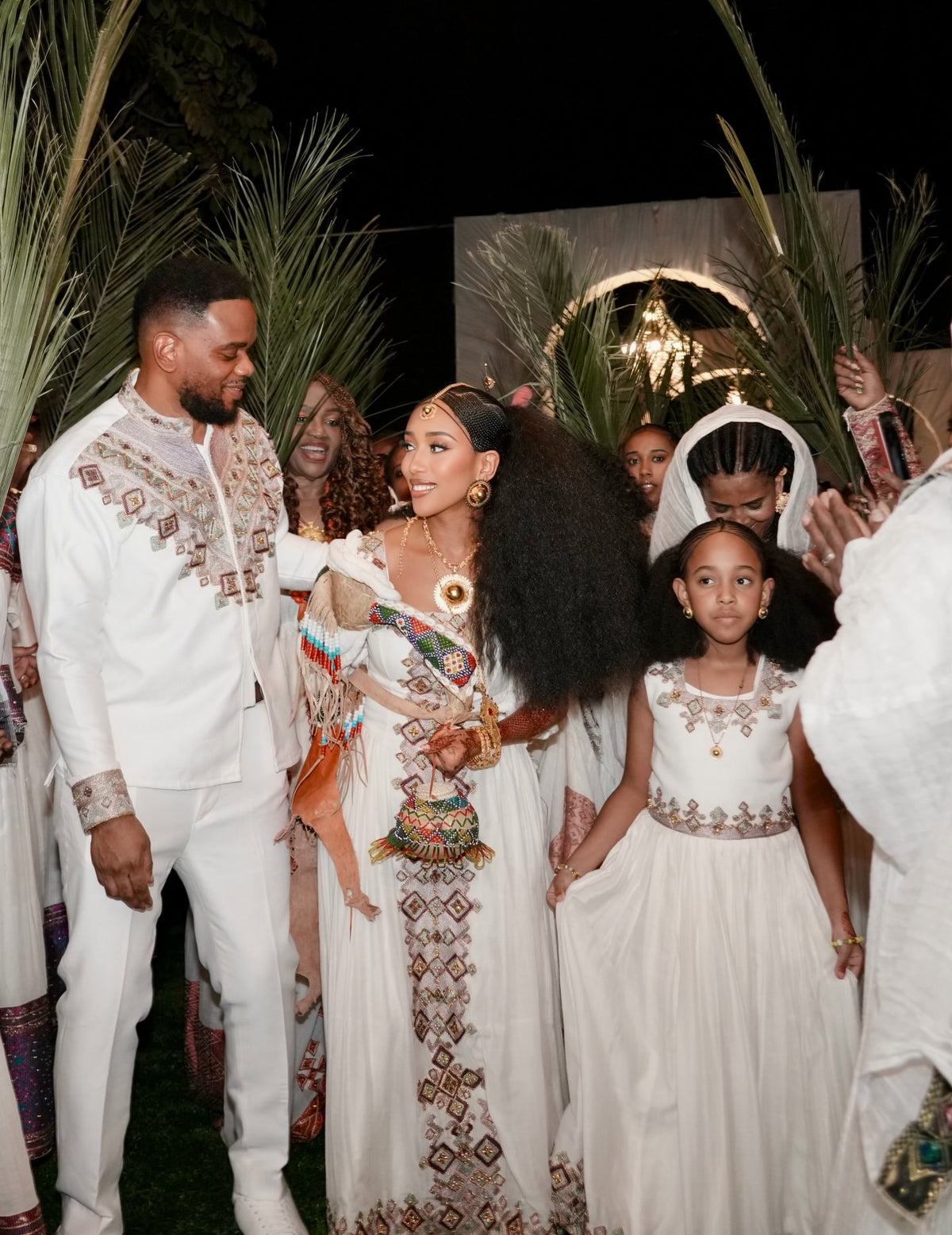 A festive nighttime celebration with a man and woman in ornate white embroidered traditional Habesha Kemis, surrounded by children and palm leaves. The woman wears a long white dress with colorful geometric embroidery and gold jewelry, and the man wears a matching white embroidered suit.