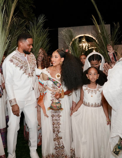 A festive nighttime celebration with a man and woman in ornate white embroidered traditional Habesha Kemis, surrounded by children and palm leaves. The woman wears a long white dress with colorful geometric embroidery and gold jewelry, and the man wears a matching white embroidered suit.