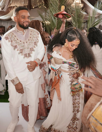 A man and a woman in ornate traditional ethnic Habesha Kemis dancing at an event. The man wears a white embroidered tunic and trousers, while the woman wears a white embellished dress with colorful patterned details and a sash.