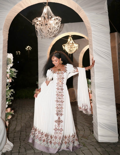 A woman wearing a white embroidered traditional Habesha Kemis with a matching dupatta, posing under draped white arches with hanging chandeliers at night.