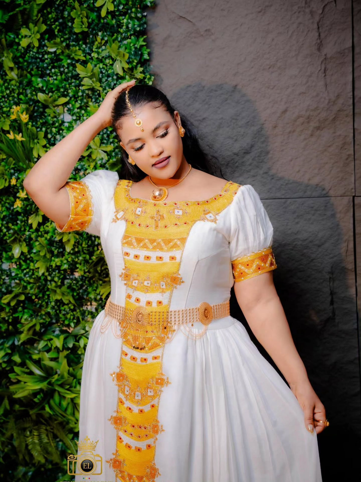 A woman wearing a white Habesha kemis with yellow and gold traditional embroidery, standing against a split background of green foliage and a dark gray wall.