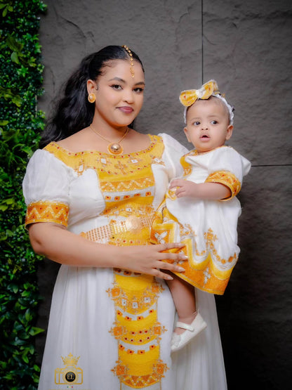 A woman and a baby wearing matching white and yellow traditional kemis , posed together against a dark textured wall with green foliage on the left.