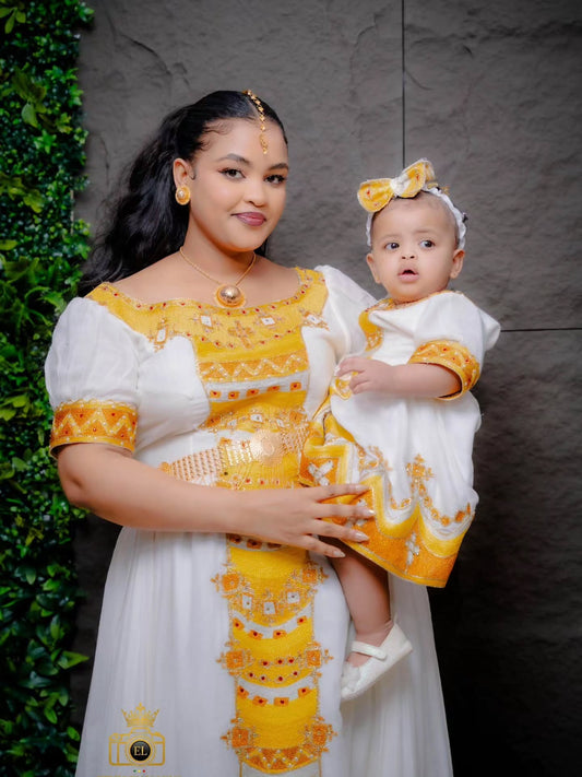 A woman and a baby wearing matching white and yellow traditional kemis , posed together against a dark textured wall with green foliage on the left.