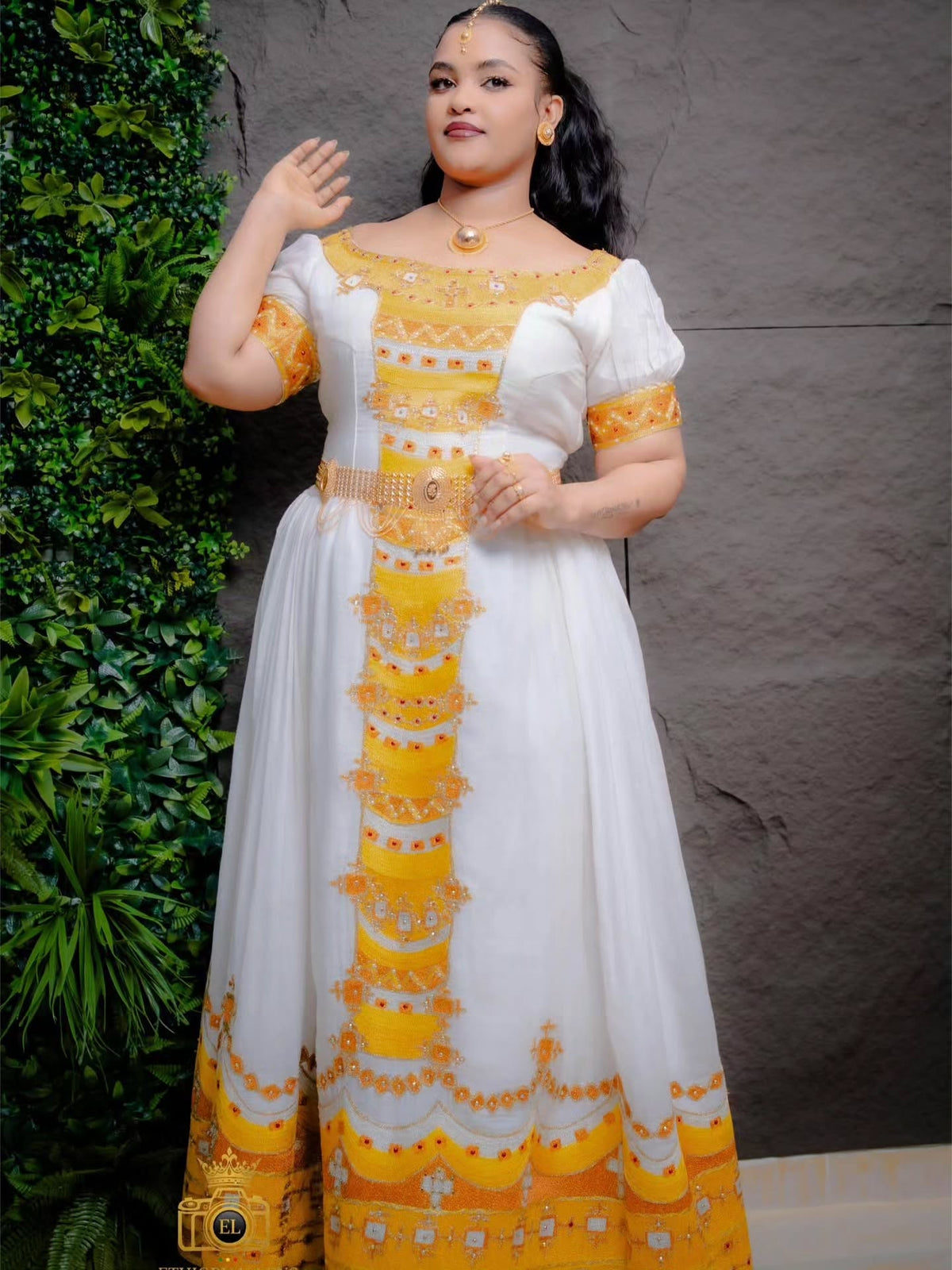 A woman wearing a white and yellow embroidered kemis with puff sleeves, standing against a textured gray wall with green foliage on the left.