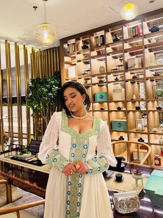 A woman standing in an elegant indoor setting wearing a white Habesha kemis with light blue and yellow embroidery along the neckline, cuffs, and front panel