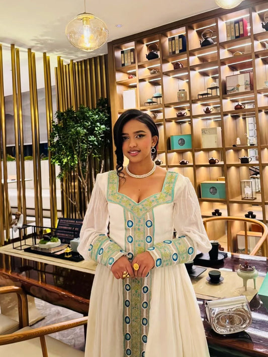 A woman standing in an elegant indoor setting wearing a white Habesha kemis with light blue and yellow embroidery along the neckline, cuffs, and front panel