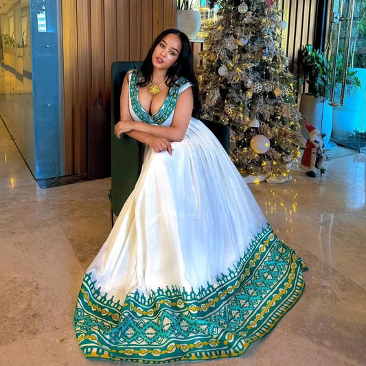 A woman wearing a white and teal traditional Indian  Habesha Kemis  with a heavily embroidered border, posing in a decorated indoor lobby next to a Christmas tree.
