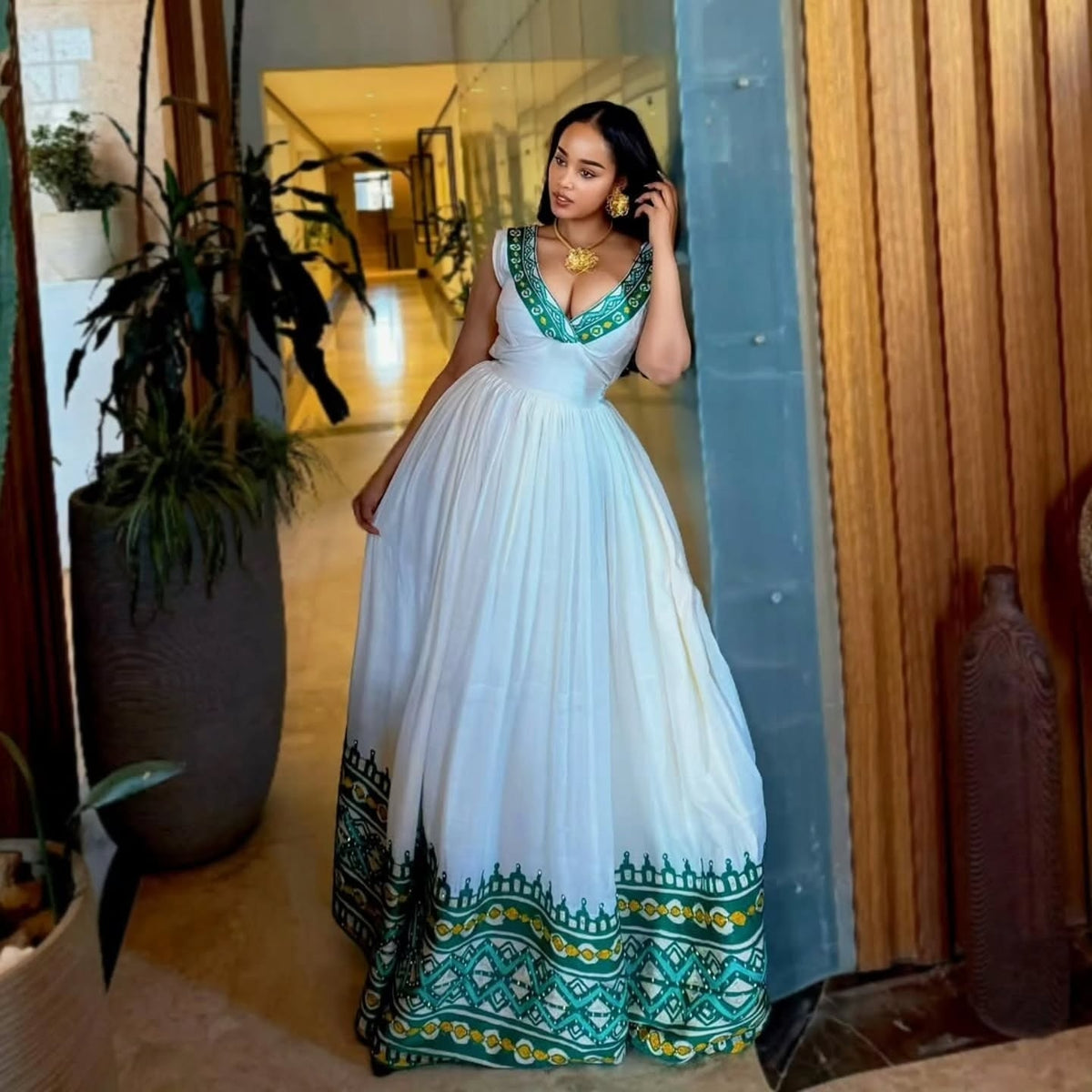 Woman in a white  Habesha Kemis  with green and blue patterns standing indoors.