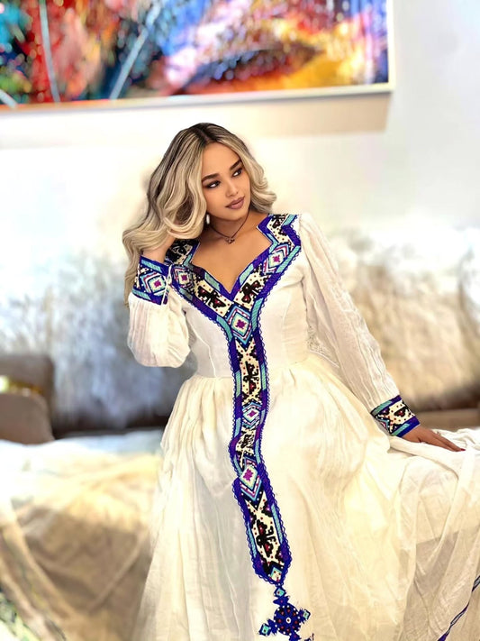  smiling woman sitting down wearing a white Habesha dress with intricate dark blue, light blue, and yellow embroidery along the deep V-neckline, front panel, and cuffs