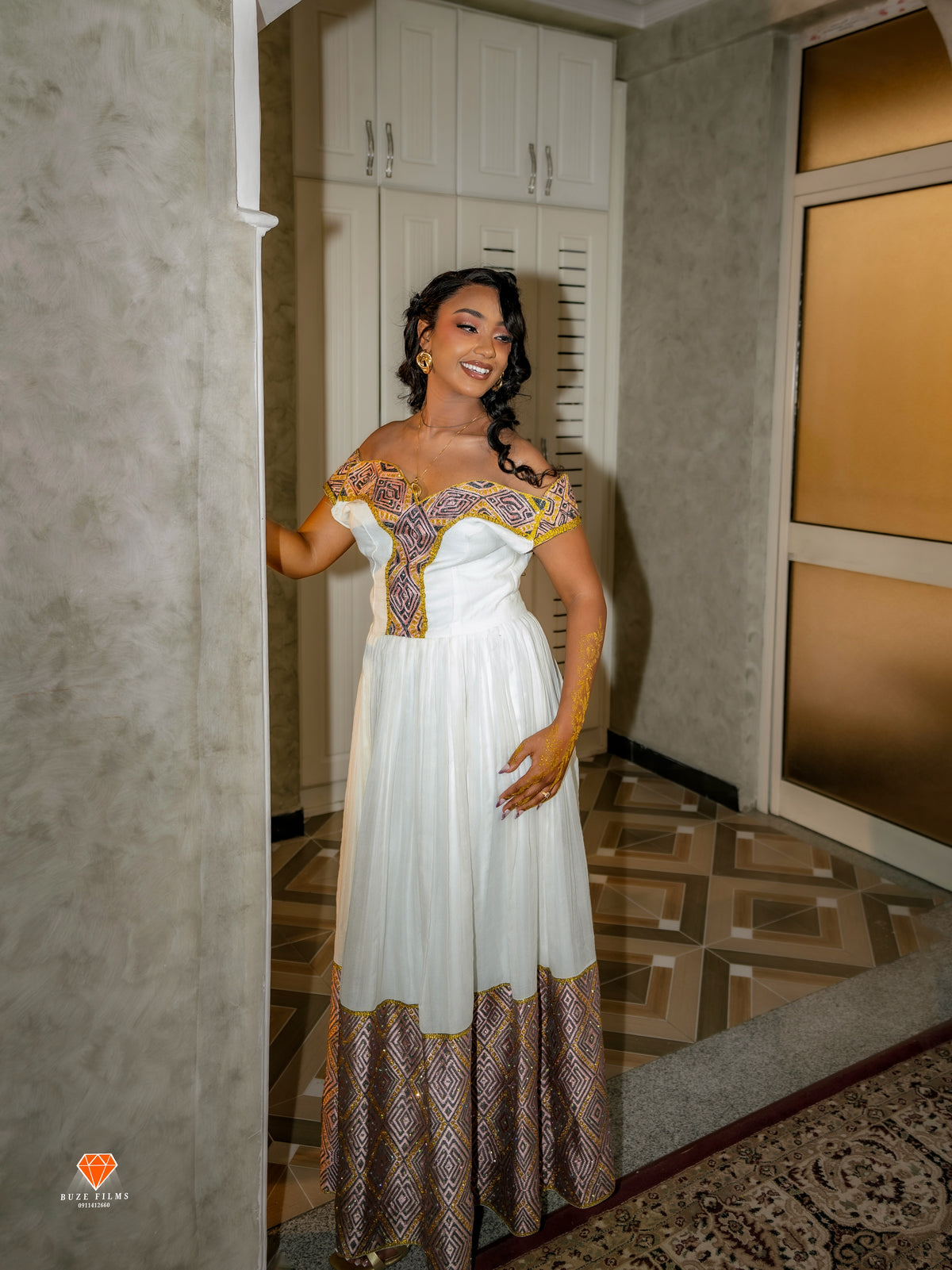 A woman models a white off-the-shoulder maxi Habesha Dress with gold and purple patterned embroidery and a matching patterned hem, standing in an indoor hallway with textured walls and patterned flooring.