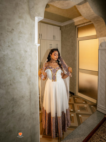 A woman wearing a white A-line Habesha Dress with ornate gold embroidery on the bodice and a patterned brown tulle hem, posing in a doorway of a textured interior hallway.