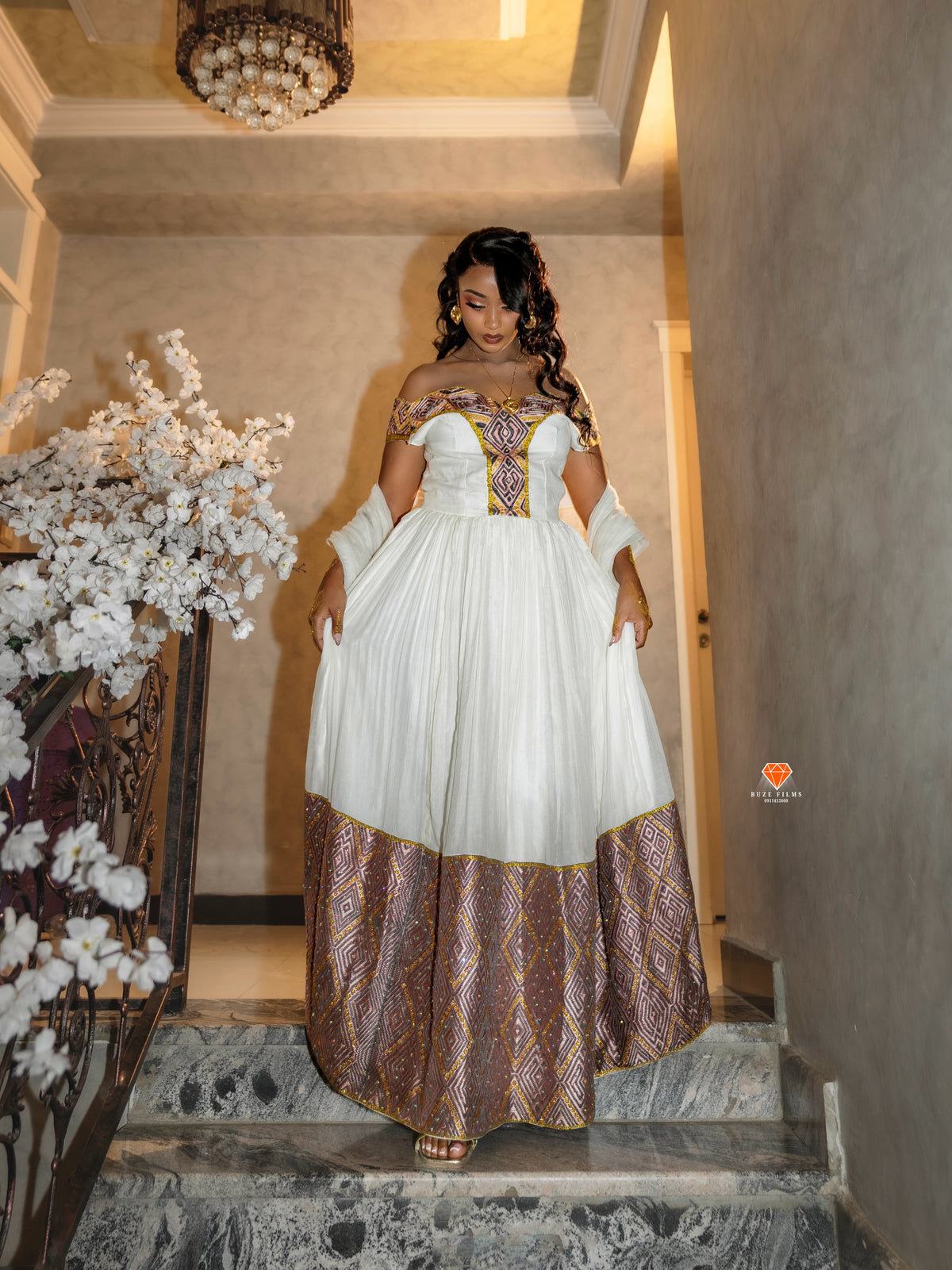 A woman wearing a traditional off-the-shoulder white Habesha Dress with gold embroidery and a brown patterned skirt, standing on stone stairs in an indoor setting.