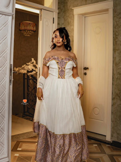 A woman wearing a white off-the-shoulder Habesha Dress with ornate purple and gold embroidery on the bodice and a matching patterned skirt, standing in an indoor hallway with cream-colored doors.