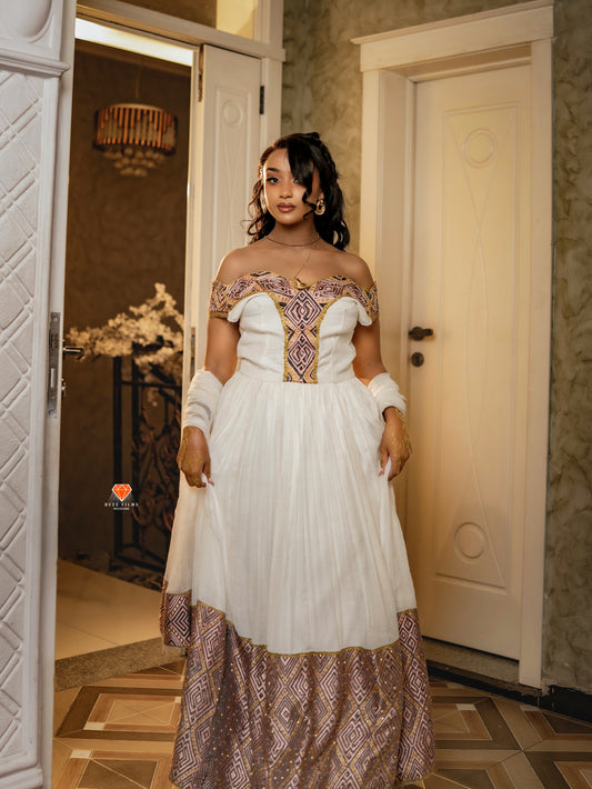 A woman wearing a white off-the-shoulder Habesha Dress with ornate purple and gold embroidery on the bodice and a matching patterned skirt, standing in an indoor hallway with cream-colored doors.