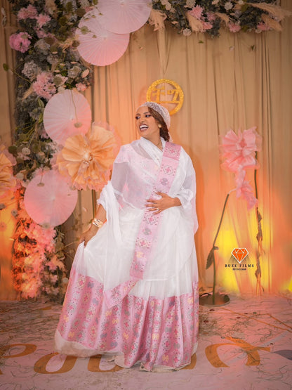Woman in a floral habesha wedding dress standing in front of a decorated backdrop with pink and orange balloons.