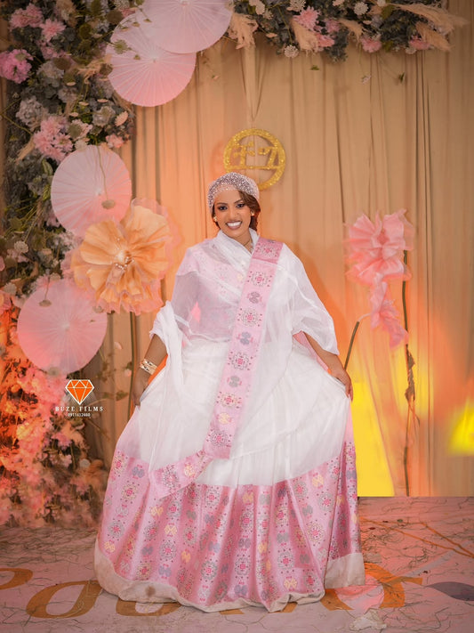 Woman in a white and pink traditional habesha wedding dress standing in front of a decorated backdrop with balloons and flowers.