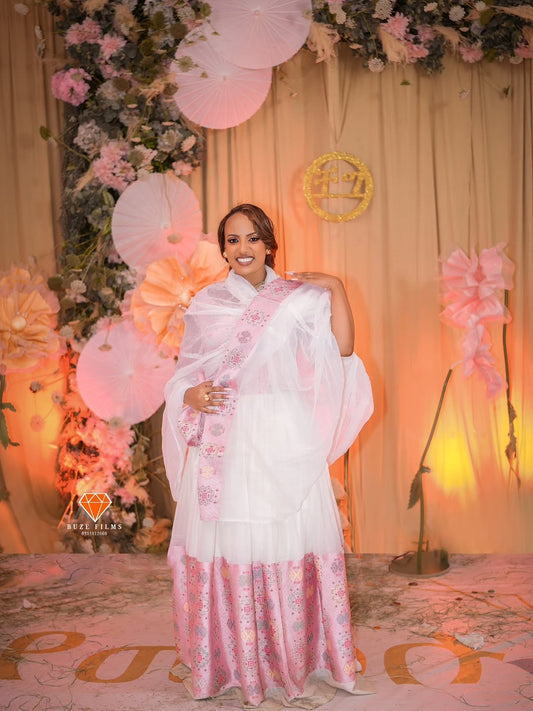Woman in a white habesha wedding dress with pink patterns standing in front of floral decorations and balloons.