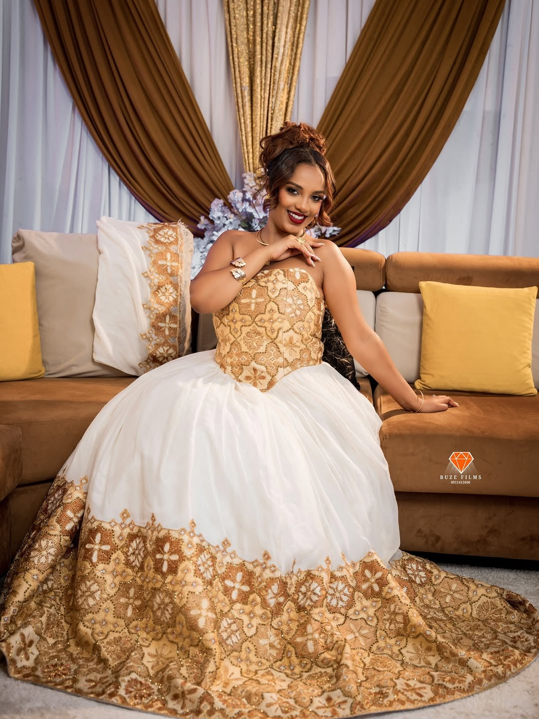 Woman in a gold and white Habesha Kemis sitting on a couch with decorative curtains and pillows.