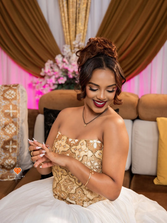 Woman in a gold and white Habesha Kemis sitting on a couch with decorative background
