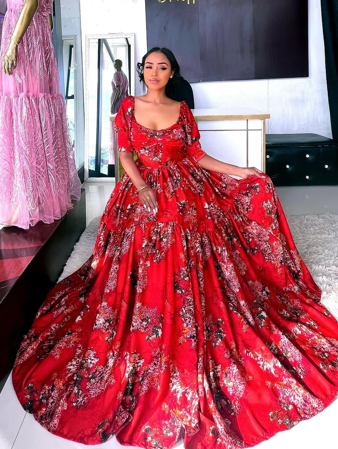 Woman wearing a red floral Shiffon Habesha in a room with a mirror and pink dress in the background.