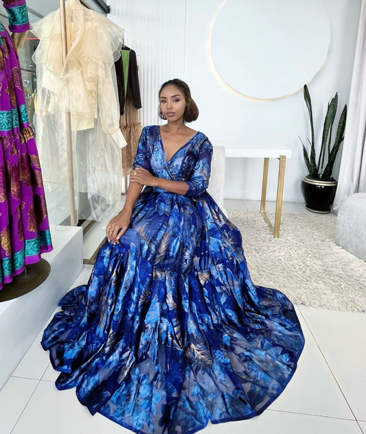 A woman wearing a voluminous royal blue ball habesha chiffion with an abstract watercolor pattern, seated in a bright boutique setting.