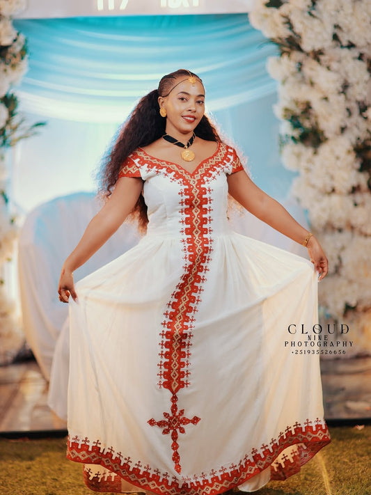 Woman in a white Traditional Dress with red embroidery standing in front of floral decorations.
