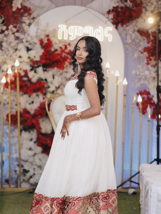 Woman in a white  Ethiopian Traditional Dress with red patterns standing in front of a decorated floral arch.