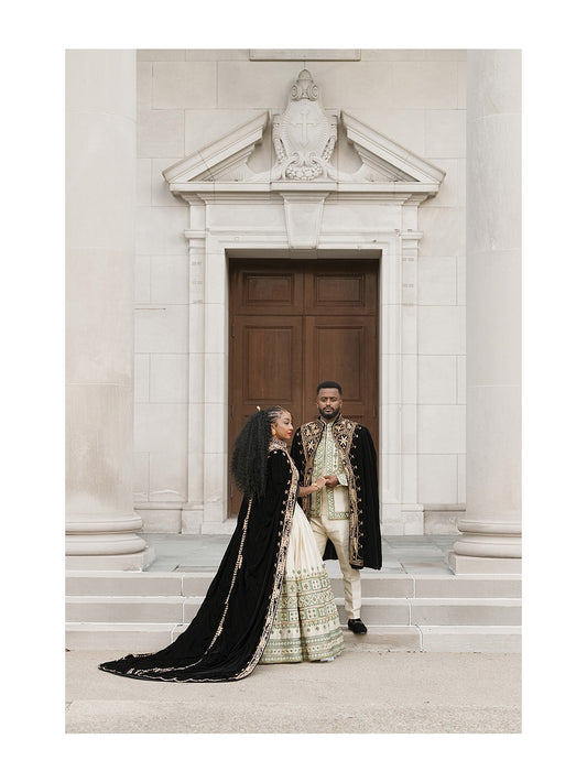 A couple in traditional Green Habesha matching couple set posing in front of a grand stone building entrance. The woman wears a black velvet bridal gown with gold embroidery and a long train, while the man wears a cream and gold embroidered sherwani with a black cape.