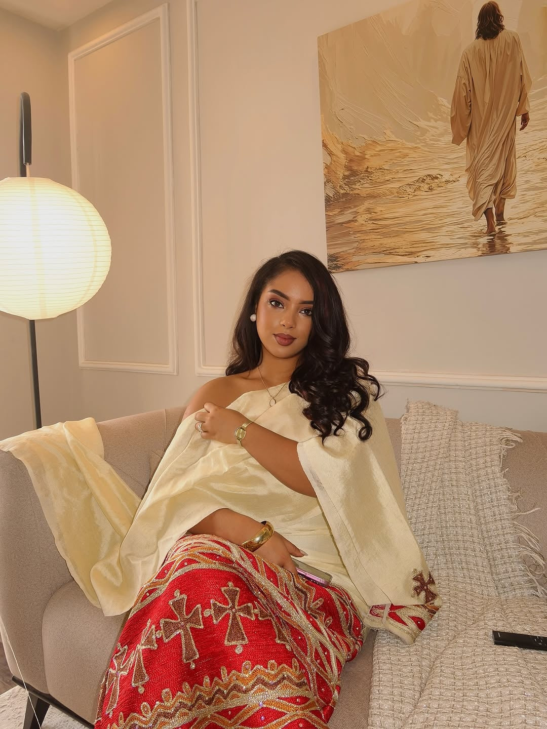 Woman in a white and red traditional outfit sitting on a couch in a room with a painting on the wall.