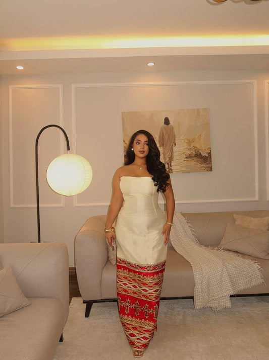 Woman in a white and red ethiopian dress standing in a living room with a couch and lamp.