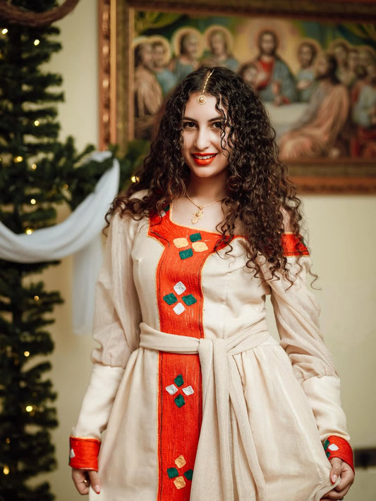 Woman wearing a Ethiopian traditional dress  with a decorative red and white pattern, standing in a room with a Christmas tree and religious icon in the background.